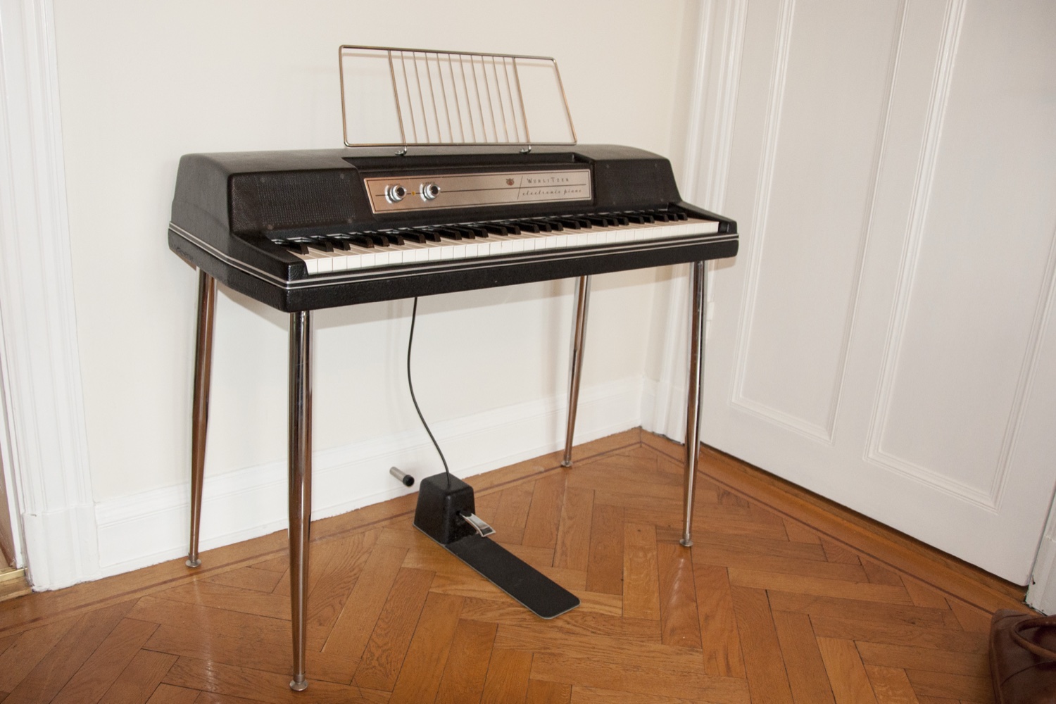 Чёрное электрическое пиано Wurlitzer на хромированных ножках с надписью electronic piano на панели, стоит у стены на паркете