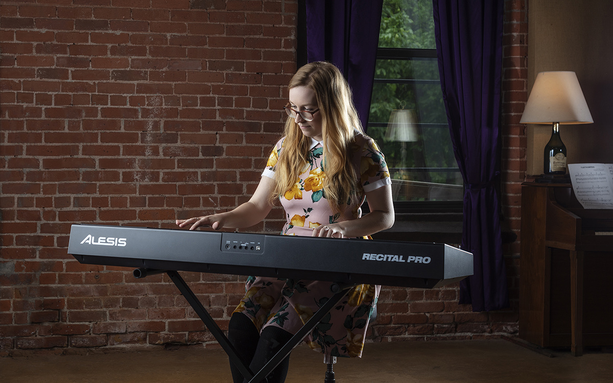 Девушка играет на чёрном Alesis Recital Pro на X-образной стойке в интерьере с кирпичной стеной