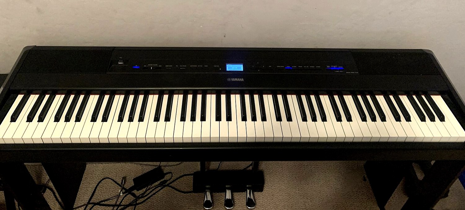 Портативное цифровое пианино Yamaha P-515 на стойке у стены: инструмент включён, на полу под ним лежит чёрный сетевой адаптер с кабелями, рядом блок из трёх педалей