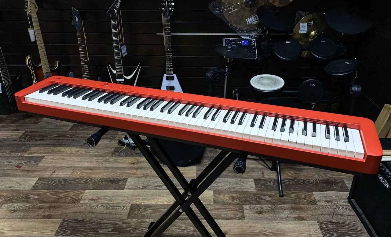 Красное цифровое пианино Casio CDP-S160 на складной X-стойке в музыкальном магазине, на стене за ним гитары
