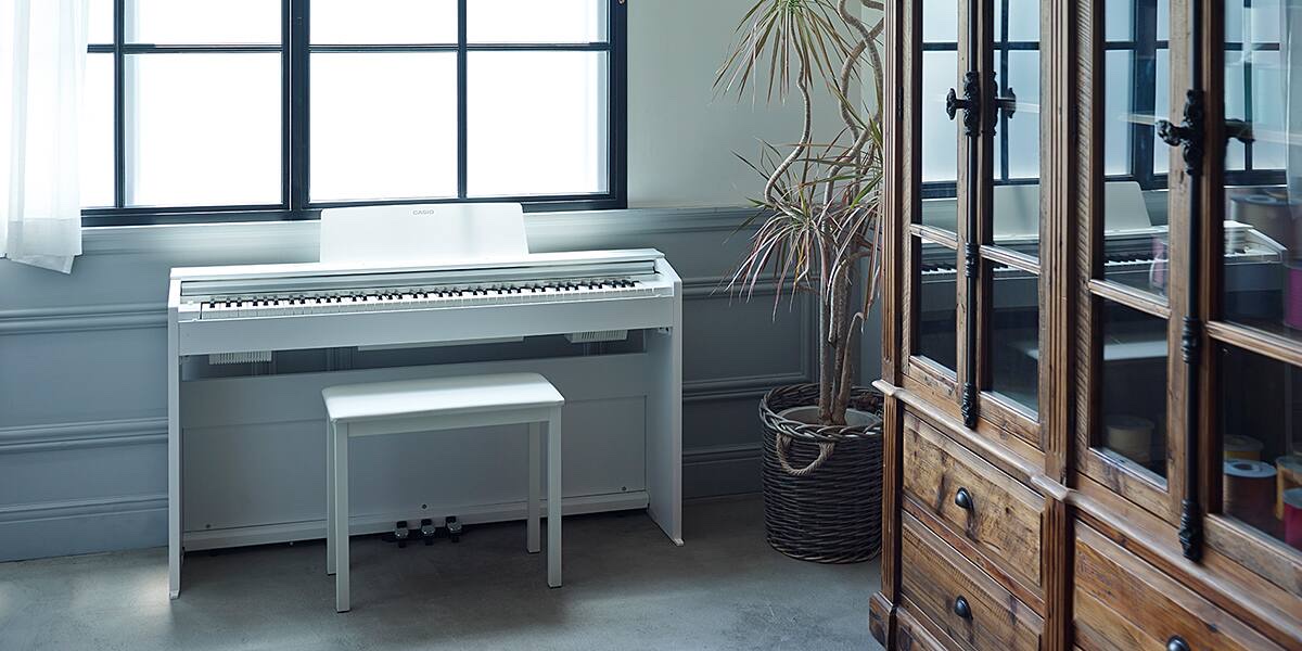 Белое корпусное цифровое пианино Casio PX-870 в исполнении WE у окна светлой комнаты: тумба с тремя педалями, белая банкетка, рядом кадка с драценой и старинный шкаф-витрина