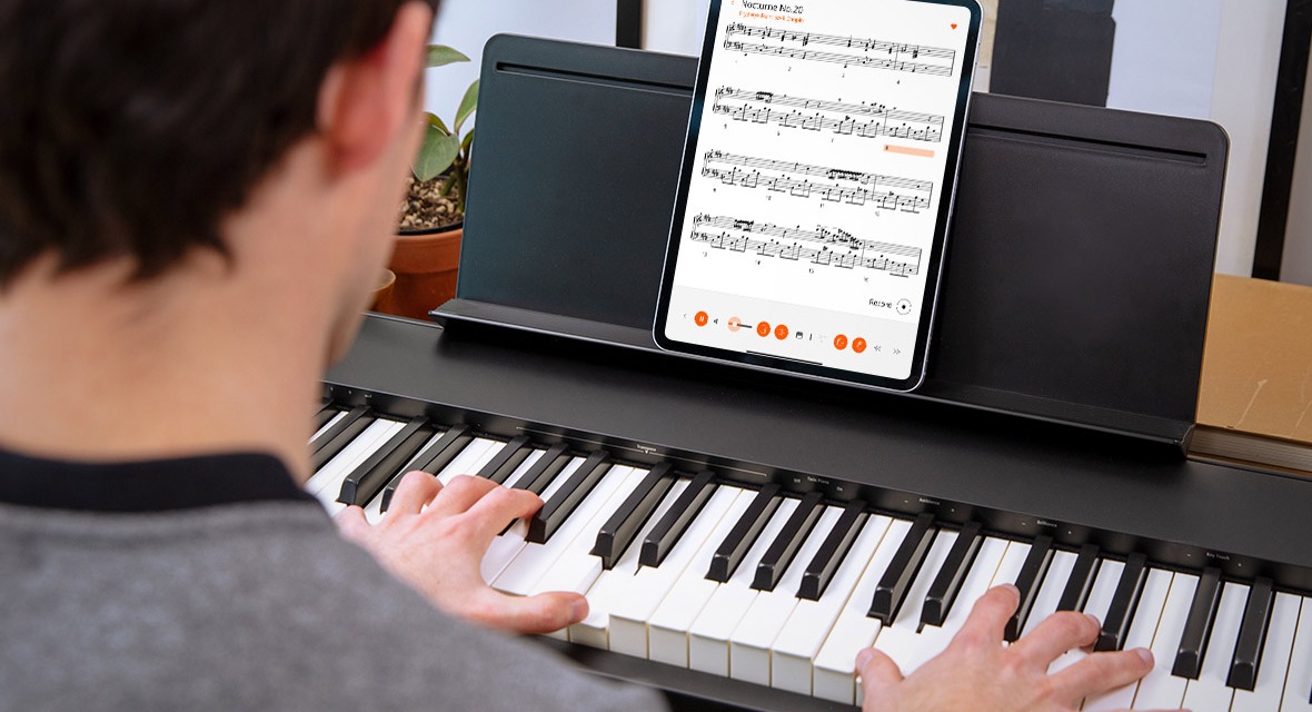 Вид из-за плеча играющего: на пюпитре чёрного цифрового пианино Roland FP-30X стоит планшет с нотами на экране, обе руки лежат на клавишах