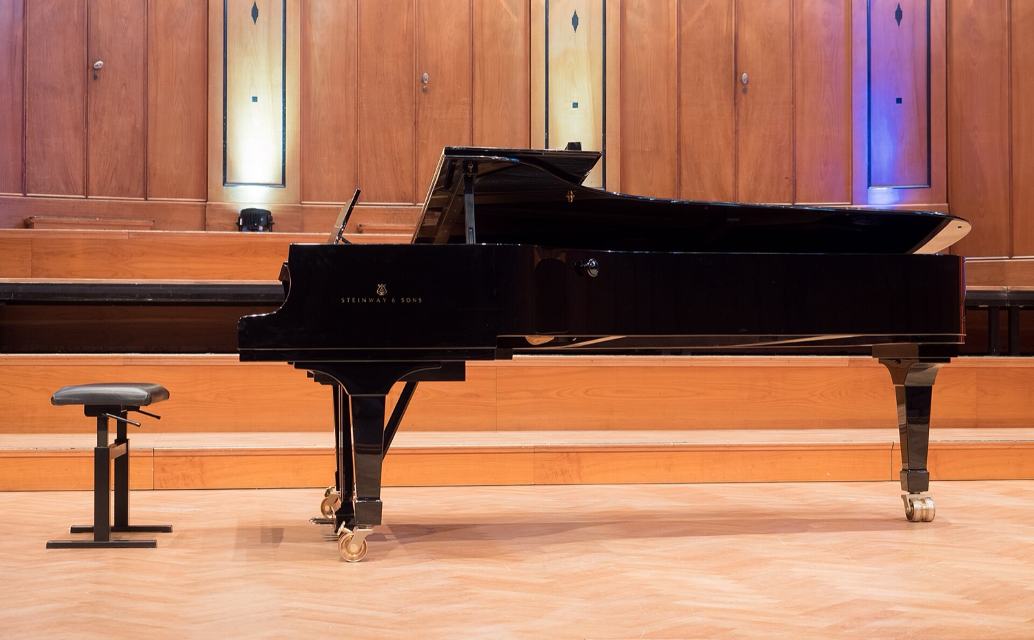 Концертный рояль Steinway & Sons чёрного полированного цвета сбоку на сцене: длинный горизонтальный крыловидный корпус на трёх ножках с колёсиками, поднятая крышка, рядом банкетка