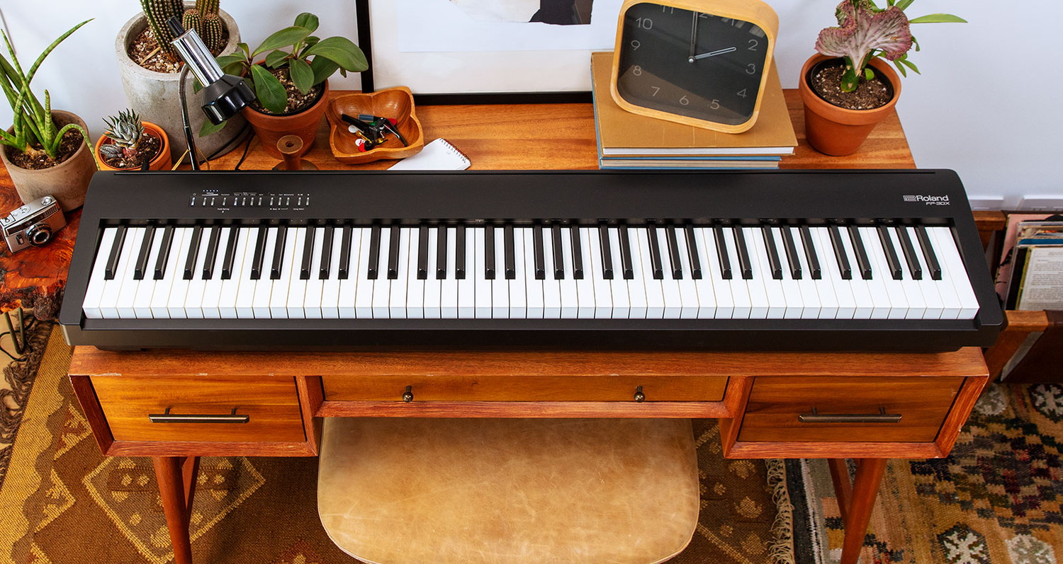 Портативное цифровое пианино Roland FP-30X на деревянном столе в комнате: над клавиатурой узкая панель с несколькими мелкими кнопками, вокруг горшки с растениями, часы и стопка книг