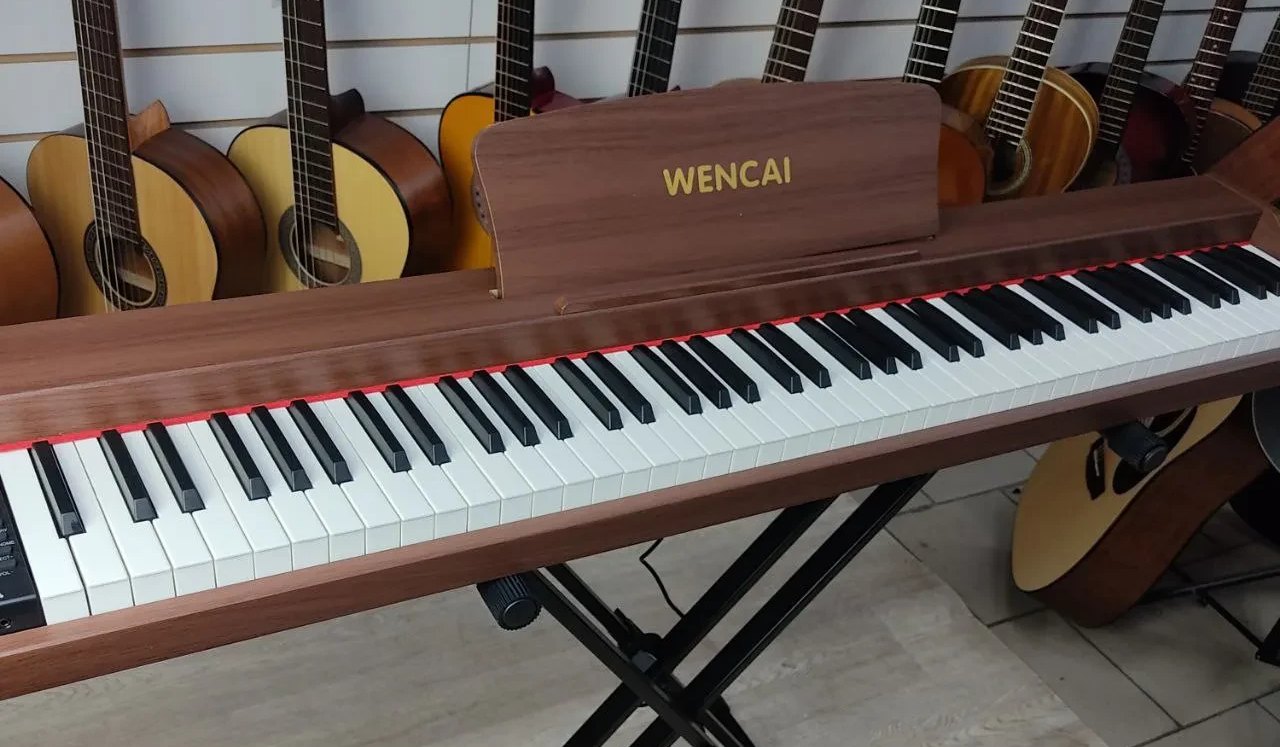 Портативное 88-клавишное цифровое пианино Wencai BL-280 в корпусе под тёмное дерево на чёрной стойке-крестовине: пюпитр с надписью WENCAI, за инструментом на стене — ряд акустических гитар музыкального магазина