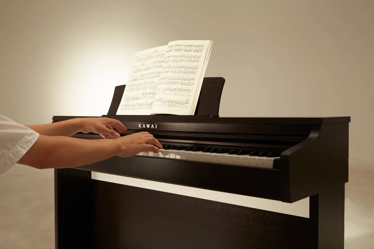 Руки на клавиатуре Kawai KDP-120 в отделке палисандр, снято сбоку: матовые пластиковые клавиши, надпись KAWAI на передней панели без кнопок и экрана, раскрытые ноты на пюпитре