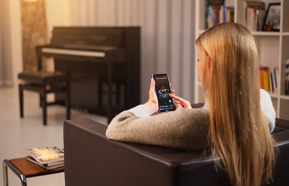 Девушка в кресле держит смартфон с открытым PianoRemote, на экране выбран тембр SK-EX Concert Grand; на заднем плане корпусное цифровое пианино Kawai