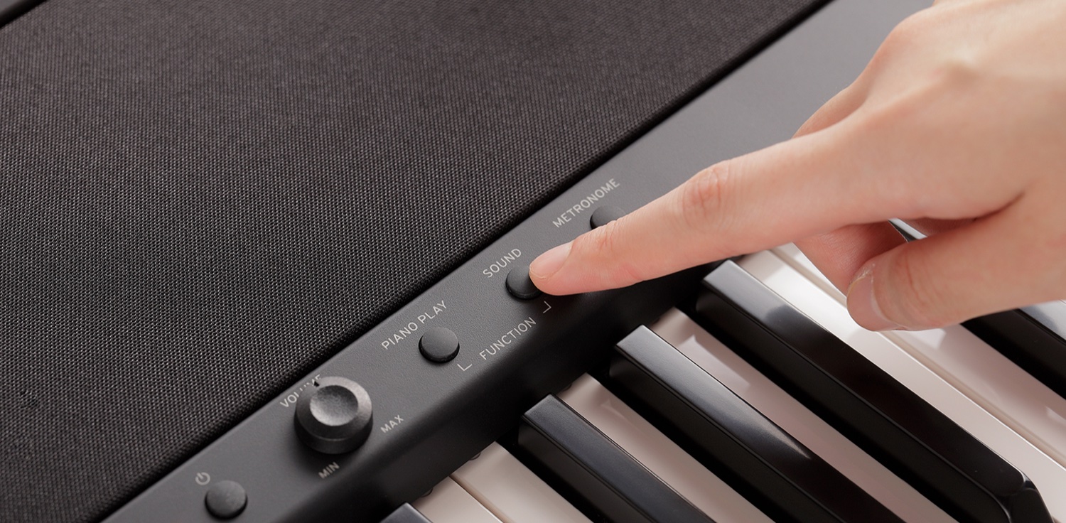 Крупный план левого края панели Korg B2: ручка громкости MIN–MAX и кнопки PIANO PLAY, SOUND и METRONOME с надписью FUNCTION, палец нажимает кнопку SOUND