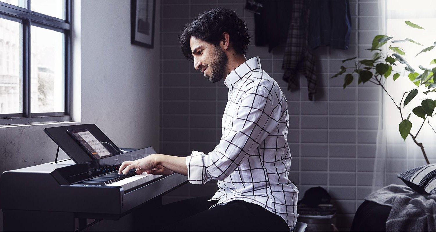 Мужчина играет на цифровом пианино Yamaha P-S500 у окна: на пюпитре стоит планшет с нотами, над клавишами горят цветные огоньки подсказки
