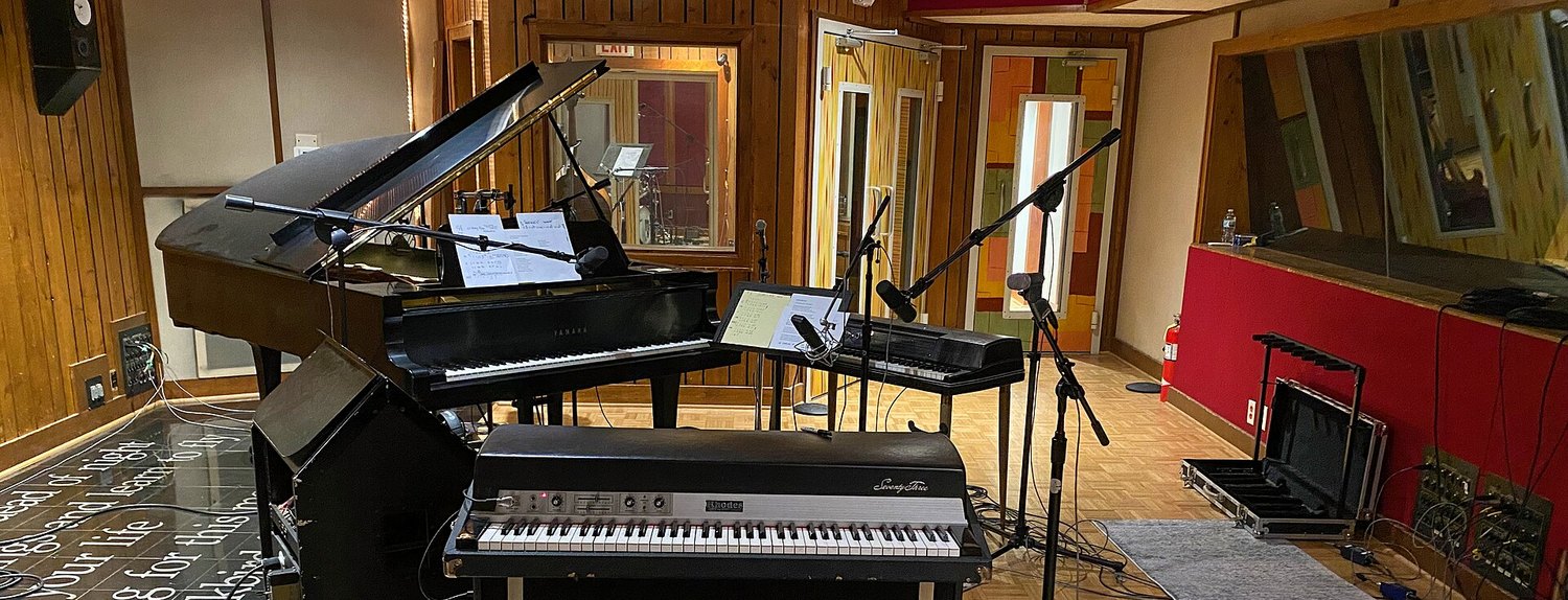 Студия звукозаписи: электропиано Fender Rhodes Seventy Three на переднем плане, за ним чёрный Wurlitzer на стойке и акустический рояль с поднятой крышкой