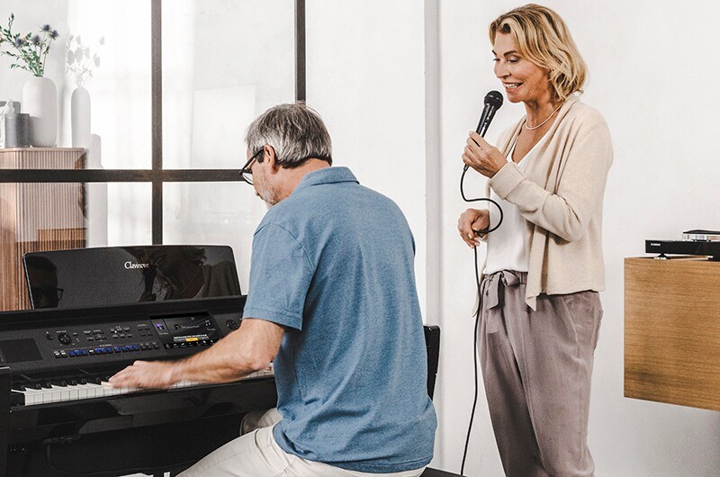 Мужчина играет на цифровом пианино Yamaha Clavinova в жилой комнате, рядом стоит женщина и поёт в проводной микрофон, кабель уходит к инструменту