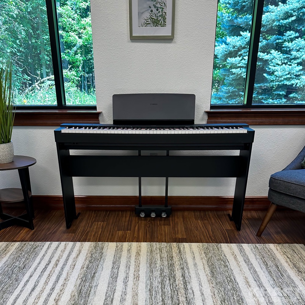 Чёрное цифровое пианино Yamaha P-225 на стойке с тремя педалями в светлой комнате у окна